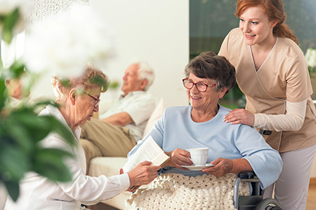 Two senior pensioners enjoying their leisure time together inside a private nursing home. Tender caretaker in uniform standing next to them